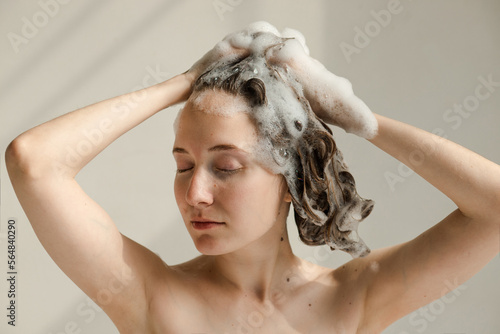 Young woman with hands touching her wet, soapy hair with shampoo foam