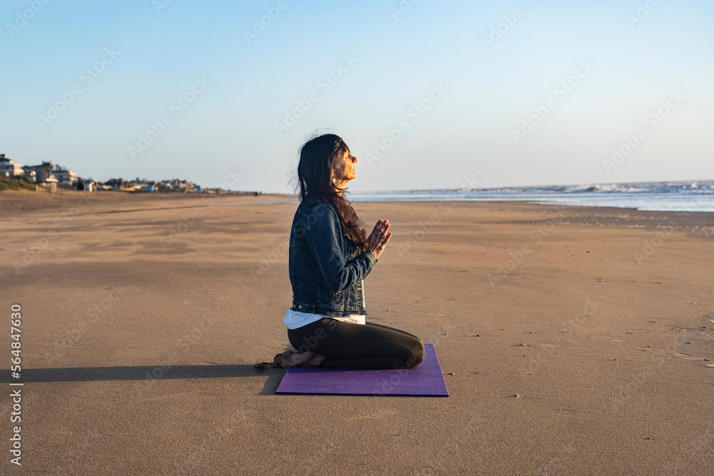Fototapeta premium Side view of a woman sitting on the sand while meditating at sunrise