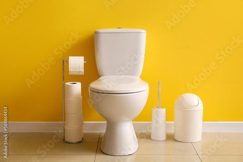 Ceramic toilet bowl, paper rolls, bin and brush near yellow wall