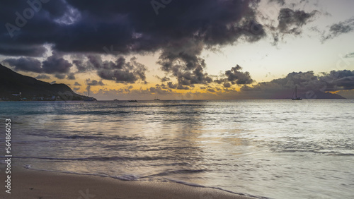 Twilight on the beach of a tropical island. Purple clouds in the sky  highlighted in orange.  Reflection on the ocean. Waves are spreading across the sand. Yachts and boats on the horizon. Seychelles