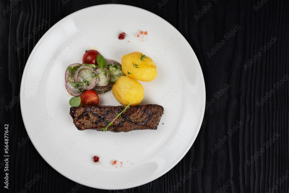 Grilled beef skirt steak meat with potato and vegetables on white plate on wooden black background