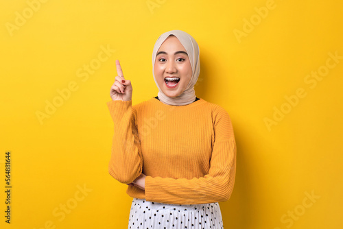 Excited beautiful Asian girl wearing hijab having creative idea, pointing finger up at copy space isolated on yellow background photo