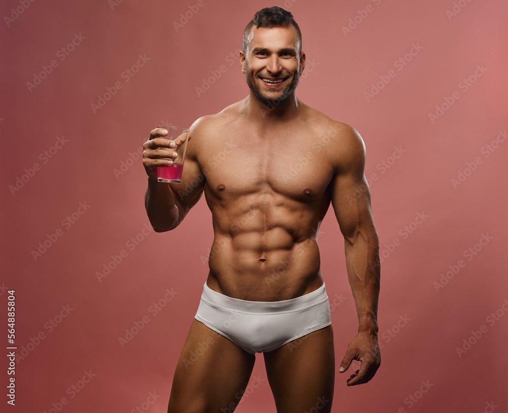 Muscular handsome man smiling and holding glass with liquid at pink background. Male fitness model posing in studio. Caucasian hunk with six pack abs. Muscular handsome man smiling and holding glass with liquid at pink background. Male fitness model posing in studio. Caucasian hunk with six pack abs.