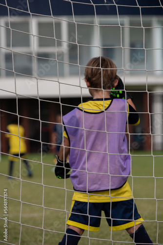 Close-up of football goal net, seeing a young soccer goalie goalkeeper during the match. Youth Soccer game on a sunny summer school tournament Day. Football match going on in a background.