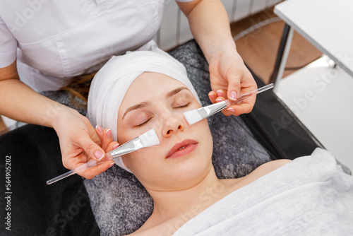 Woman getting facial peeling by cosmetologist. Beautician applying clay face mask on woman face. Cosmetologist applying mask on clients face in spa salon. Spa cosmetologist applying facial mask.