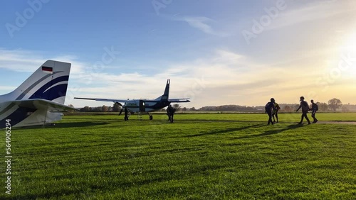 Wide shot of a few parachuters getting ready and walking to the plane on the ground. Static camera, nice afternoon sun.
