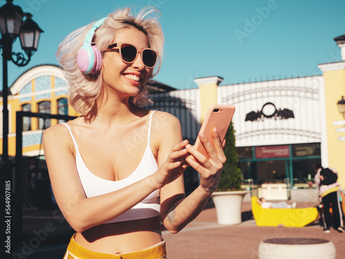 Young beautiful smiling hipster woman. Sexy carefree model posing in the street at sunset. Positive blond female enjoying listening music in wireless headphones outdoors. Holding smartphone