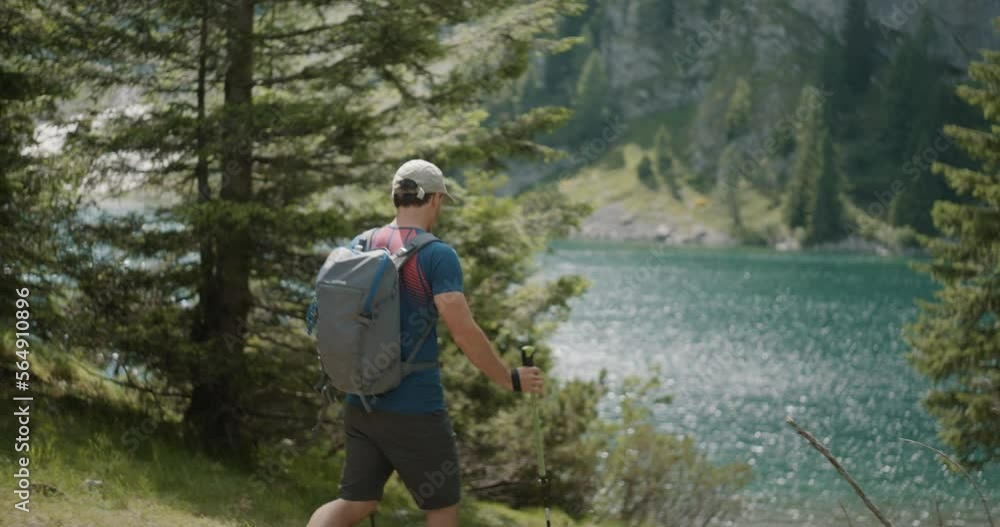 Hiker walking with his hiking poles towards the mountain lake, path surrounded by conifers, water glistening. Hiker walking with his hiking poles towards the mountain lake, path surrounded by conifers, water glistening.