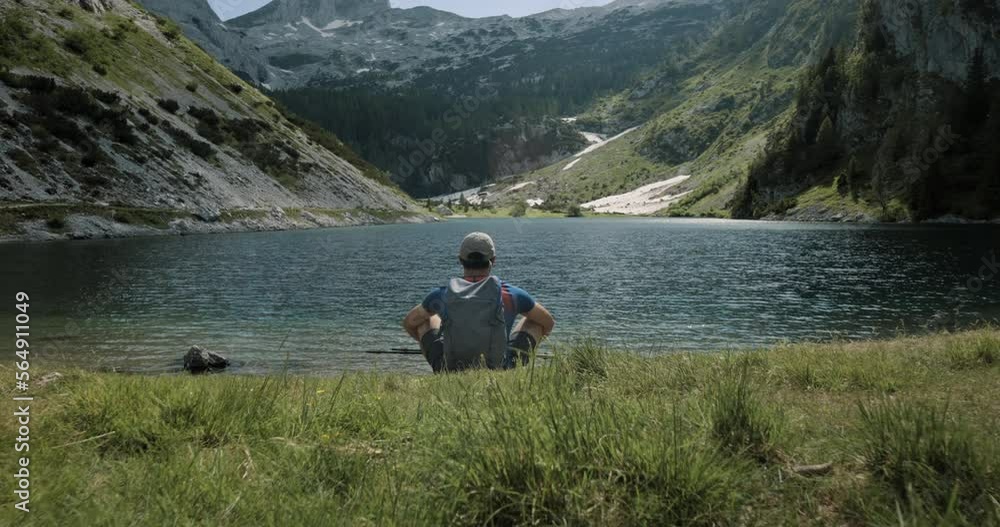 Camera tracking forward towards the lake where a hiker is sitting and looking at the valley. Mountains in the background. Camera tracking forward towards the lake where a hiker is sitting and looking at the valley. Mountains in the background.