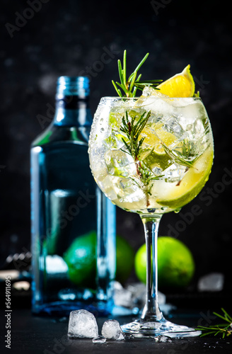 Gin tonic cocktail with dry gin, rosemary, tonic, lime and ice cubes in wine glass. Black bar counter background, bar tools