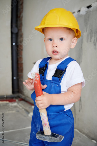 Adorable child construction worker holding hammer building repair tool and looking at camera. Cute kid wearing safety helmet and work overalls while posing at home during renovation.