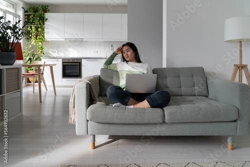 Thoughtful pensive young Asian woman freelancer looking aside while sitting on sofa with laptop, losing focus while working remotely from home. Freelance procrastination, downsides of remote work