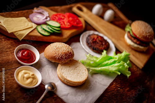 Cooking hamburger or cheeseburger. Different ingredients for a classic hamburger. Grilled meat, vegetables, greens, sauces near a sesame bun. Wooden background. Hamburger day.