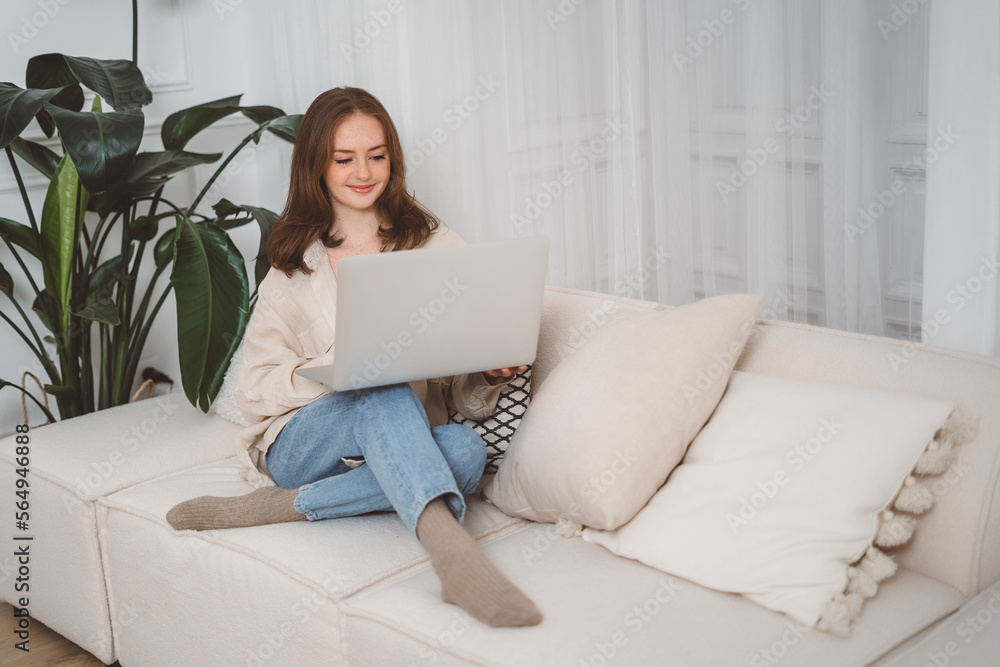 Beautiful cheerful redhead girl using silver laptop while sitting on sofa in living room at home Beautiful cheerful redhead girl using silver laptop while sitting on sofa in living room at home