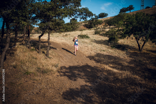 An attractive blonde girl in striped shorts and a blue t-shirt walks along a path in the hills, around the trees, on a bright sunny day. © Ihor