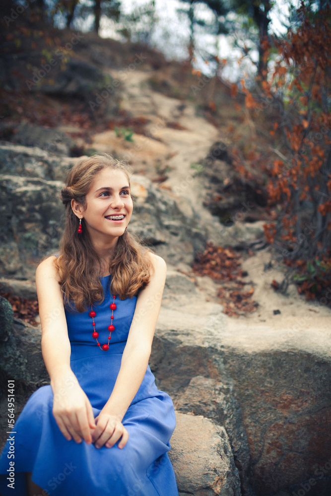 A positive beautiful young girl in a blue dress is resting in the mountains around beautiful nature, picturesque stones and trees.