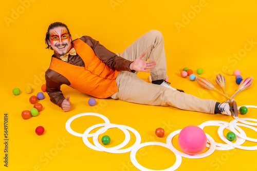 Juggler in a vest and with a painted face lying with the juggling objects on a yellow background