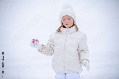 a little girl in winter white clothes in the snow holding a transparent box with candies in the shape of hearts