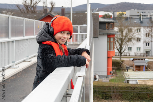 An 11-year-old girl in a red hat and headphones on a pedestrian bridge smiles and looks at the camera.