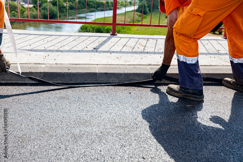 Workers apply black bitumen membrane strips for waterproofing roads and bridges to the curb of the sidewalk