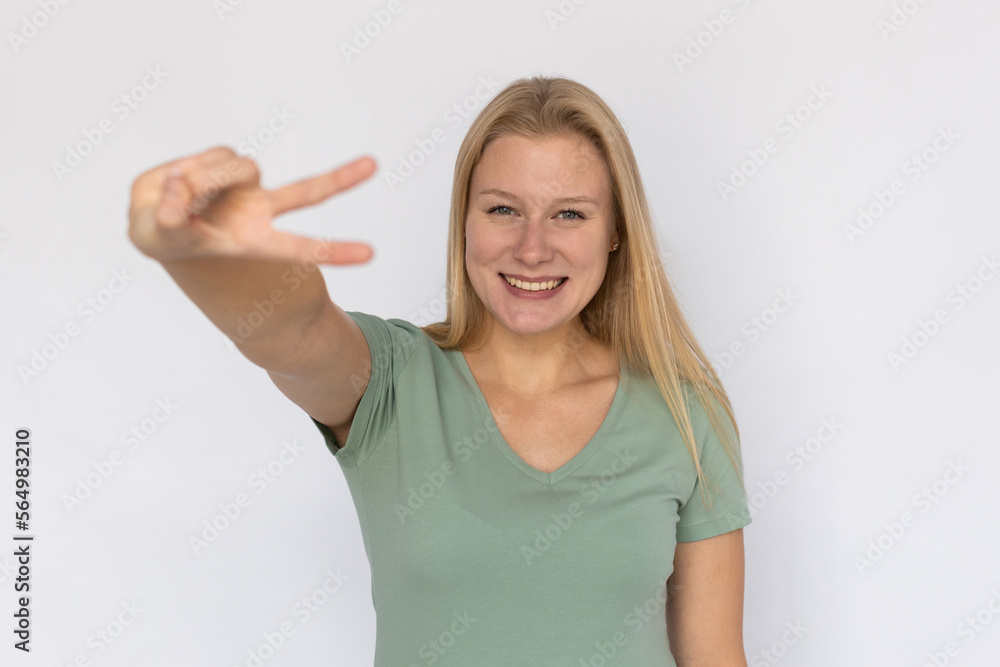 Fototapeta premium Excited young woman showing victory gesture. Portrait of happy Caucasian female model with fair hair in green T-shirt looking at camera, smiling and greeting someone. Success, optimism concept