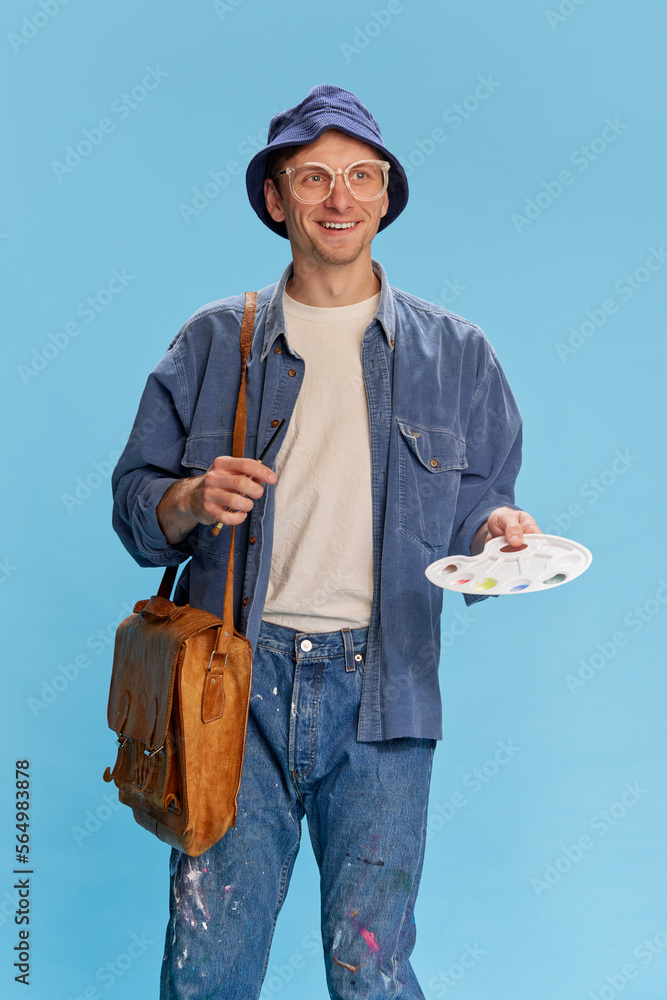 Naklejka premium Positive mood, inspired look. Portrait of young man in jeans clothes, painter, artist posing over blue studio background. Concept of emotions, facial expression, lifestyle, hobby, occupation. Ad