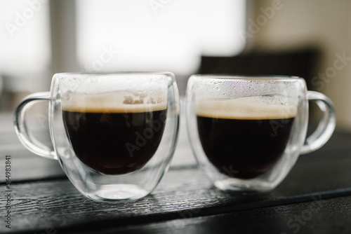 Two cups of hot black coffee on a dark wooden table. Ground or instant coffee. Delicious and strong aromatic Americano or espresso. Cups glass transparent on dark background. Side view. Copy space.