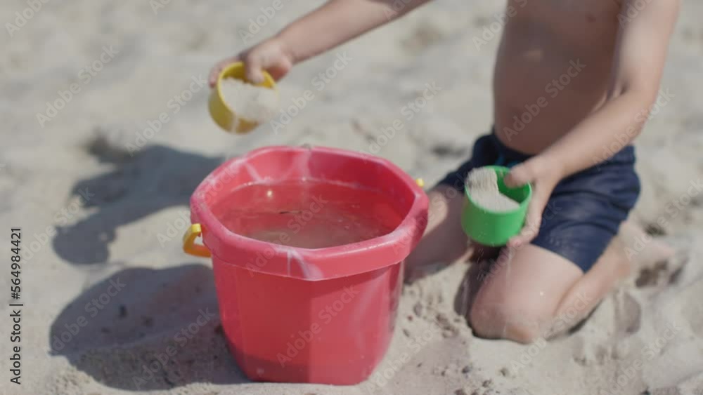 close-up child playing sand and water on beach summer sunny daytime. little boy pouring sand into bucket water exploring nature. development skills and knowledge outdoors child's kid's self education close-up child playing sand and water on beach summer sunny daytime. little boy pouring sand into bucket water exploring nature. development skills and knowledge outdoors child's kid's self education