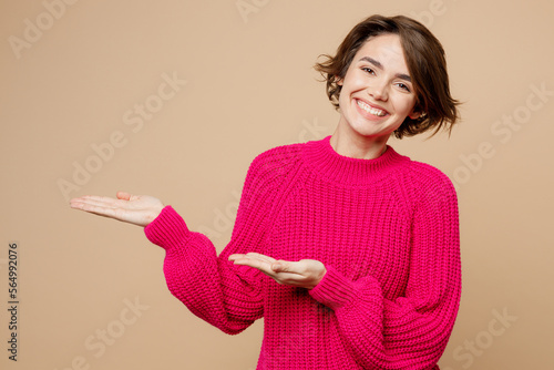 Bild auf Leinwand Young smiling woman wears pink sweater point hands arms aside indicate on workspace area copy space mock up isolated on plain pastel light beige background studio portrait