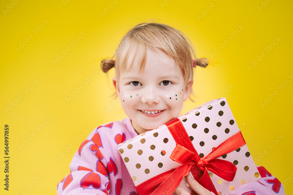 Cheerful little girl in pink sweatshirt with red hearts pattern holding gift box with red ribbon isolated over yellow background. Cheerful little girl in pink sweatshirt with red hearts pattern holding gift box with red ribbon isolated over yellow background.