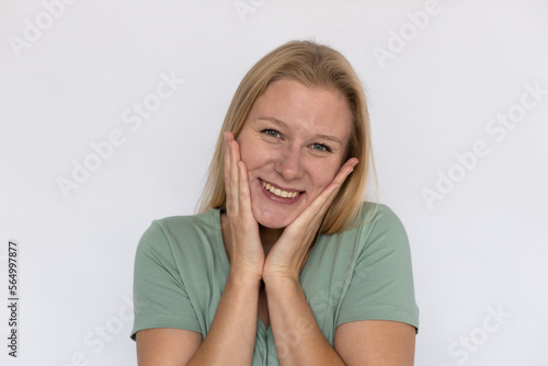 Pleased young woman touching face. Closeup of happy Caucasian female model with fair hair in green T-shirt looking at camera, smiling with hands on cheeks. Optimism, advertising concept
