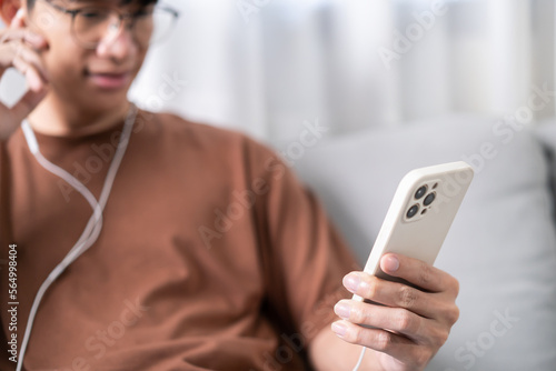 Relaxed young asian man music lover sitting listening music on earphones. Man holding mobile phone in living room listening to music with headphones and mobile phone