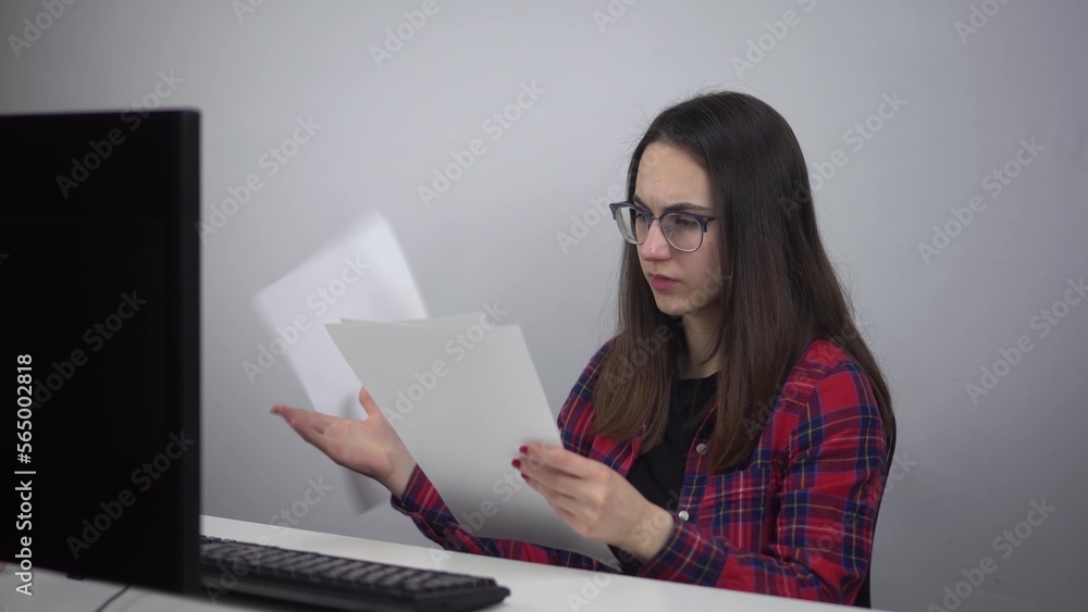 Naklejka premium A young IT lady reads documents and throws them out of anger. A woman is sitting at the workplace in the office. Girl in a red shirt and glasses.