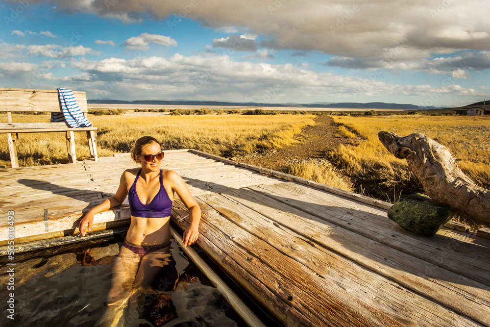 ALVORD HOT SPRINGS, OR, USA. A young woman in a bikini sits in a natural hot spring tub surrounded by a vast marshy expanse under clouds and blue sky.