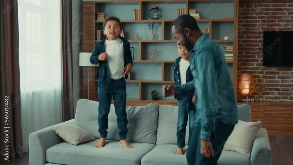 Cute active African American family dancing at home party. Ethnic adult father dad with preteen children sons kids little children dance to music at living room jumping on couch sofa having fun laugh Cute active African American family dancing at home party. Ethnic adult father dad with preteen children sons kids little children dance to music at living room jumping on couch sofa having fun laugh