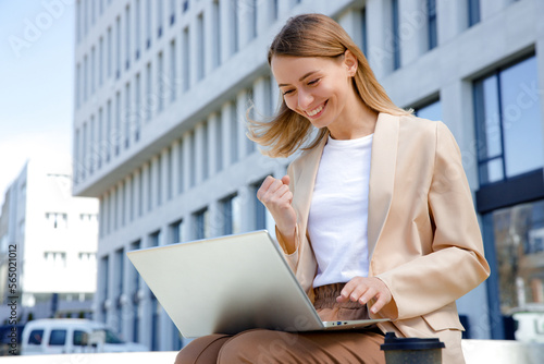 Excited businesswoman reading good news on modern laptop while sitting outdoors. Happy lady worker in suit making yes gesture with office center on background.