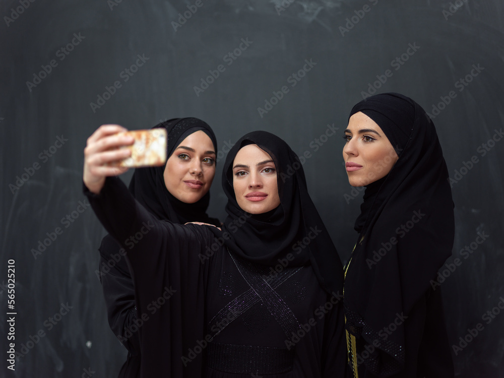 Group of young beautiful muslim women in fashionable dress with hijab using smartphone while taking selfie picture in front of black background Group of young beautiful muslim women in fashionable dress with hijab using smartphone while taking selfie picture in front of black background