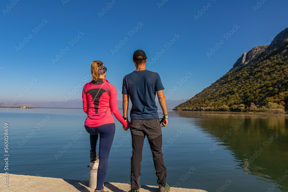 Fototapeta premium Couple standing holding hands on pier with panoramic view of Lake Skadar National Park in autumn seen from Godinje, Bar, Montenegro, Balkans, Europe. Stunning travel destination in Dinaric Alps