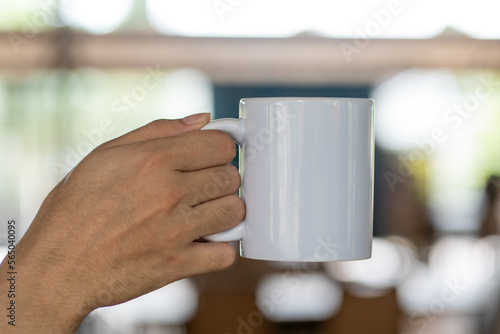 A hand holding out a white blank tea mug with out of focus outdoor cafe background