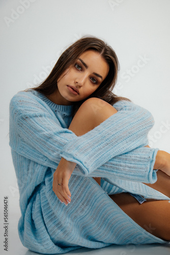 Portrait of young beauteous woman wearing long blue knitted dress, sitting on floor, hugging knees on white background.