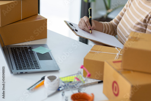 Woman starting a small business in a home office is working on a laptop to check orders from the internet and write notes in preparation for delivery.