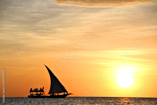 A traditional dhow boat sails through a calm and beautiful blue ocean silhouetted by the setting sun in Zanzibar, Tanzania, Africa.