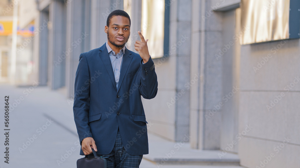Confident African American serious man employee entrepreneur businessman ethnic male guy worker manager with suitcase in business suit show number one finger hey you choosing posing in city outdoors Confident African American serious man employee entrepreneur businessman ethnic male guy worker manager with suitcase in business suit show number one finger hey you choosing posing in city outdoors