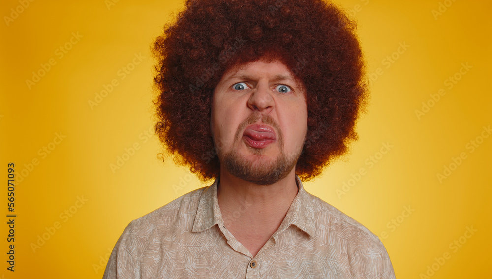 Funny joyful sincere man with lush Afro hairstyle coiffure making playful silly facial expressions and grimacing fooling around showing tongue. Young handsome hipster guy isolated on yellow background Funny joyful sincere man with lush Afro hairstyle coiffure making playful silly facial expressions and grimacing fooling around showing tongue. Young handsome hipster guy isolated on yellow background