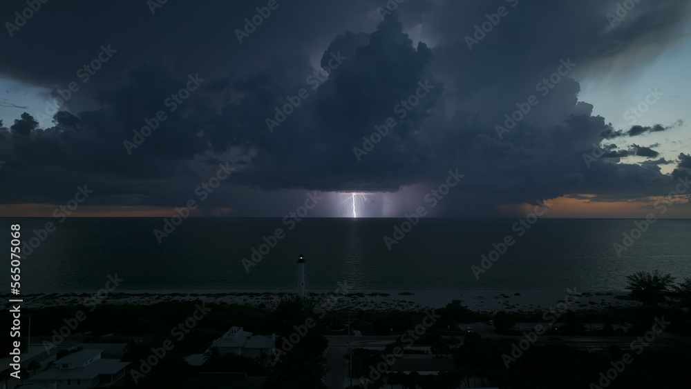 White tall lighthouse on sea shore with blinking light at stormy night for commercial vessels navigation. Thunderstorm with lightnings over ocean water posing danger for ships White tall lighthouse on sea shore with blinking light at stormy night for commercial vessels navigation. Thunderstorm with lightnings over ocean water posing danger for ships