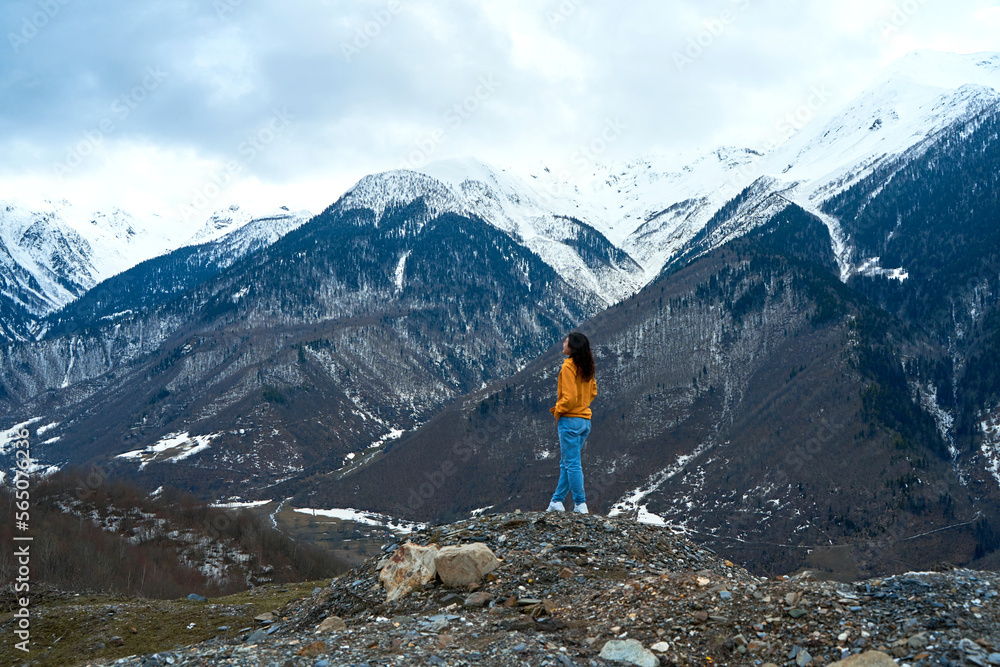 A girl in a yellow jacket and blue jeans enjoys the cold mountain scenery of snow-covered mountains