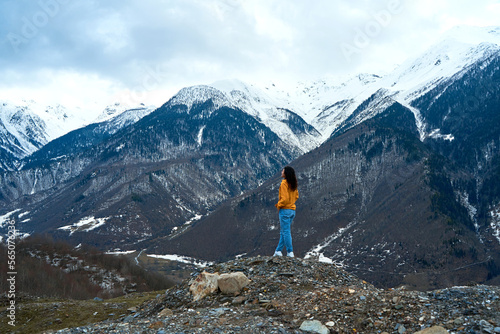 A girl in a yellow jacket and blue jeans enjoys the cold mountain scenery of snow-covered mountains