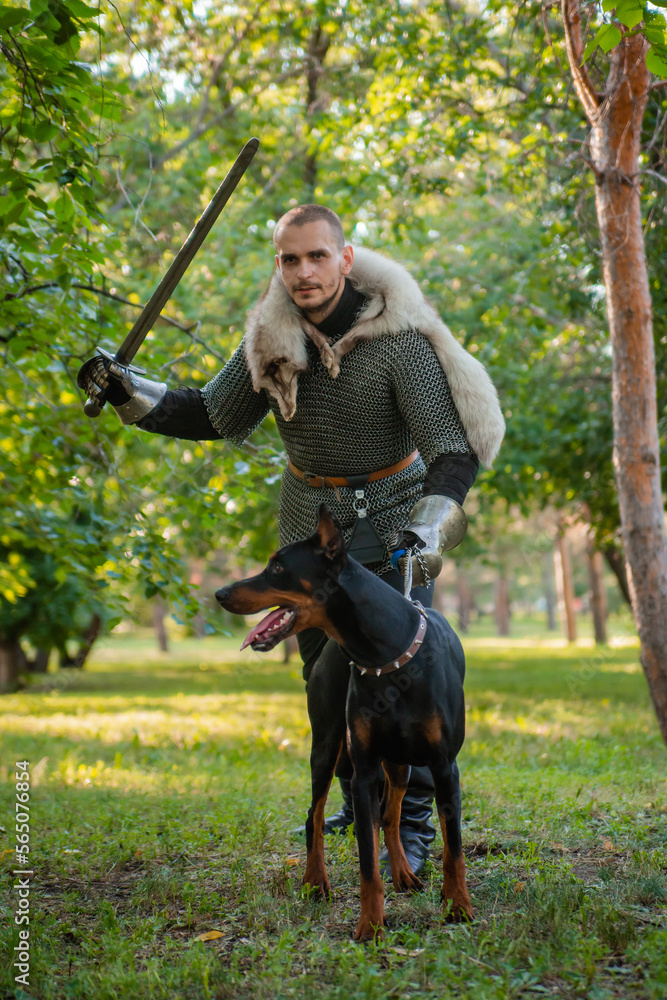 A young man in chain mail with a sword in the woods with a dog. A warrior in armor stands with a Doberman on a chain.