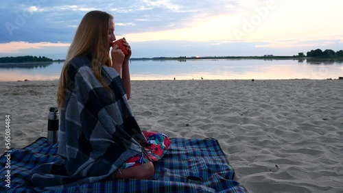 A beautiful girl sits on the beach early in the morning and admires the sun rising over the river and drinks hot coffee.