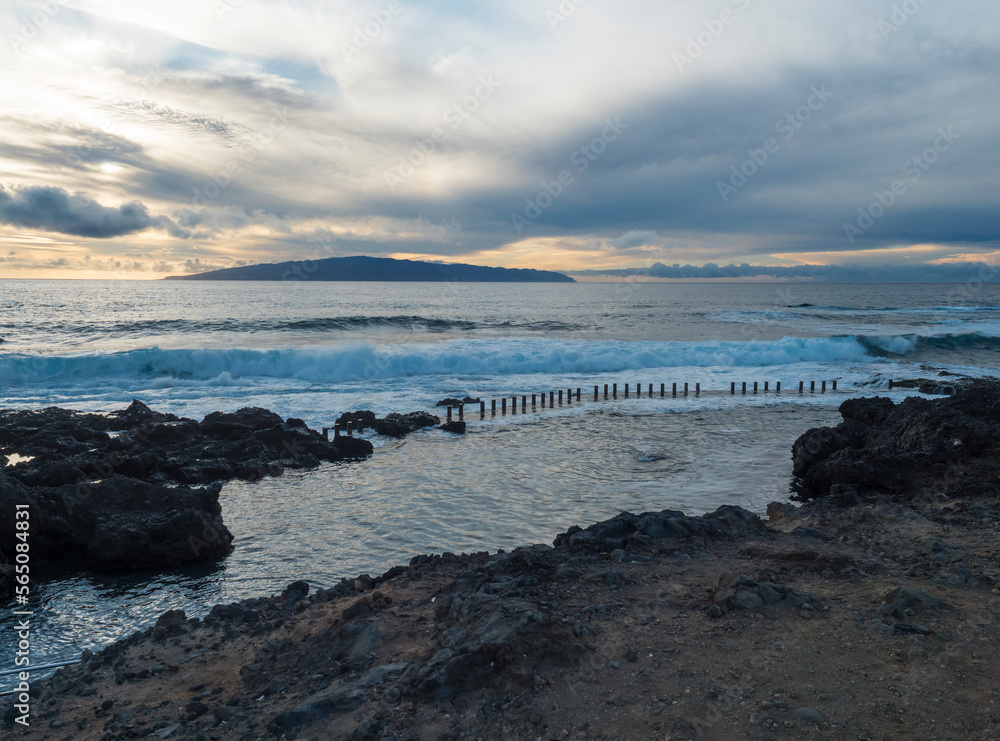 Fototapeta premium View of the natural sea rock pool on the Tenerife coast near Alcala with vawes crushing against concrete wall and black volcanic rocks. Evening sunset sky.